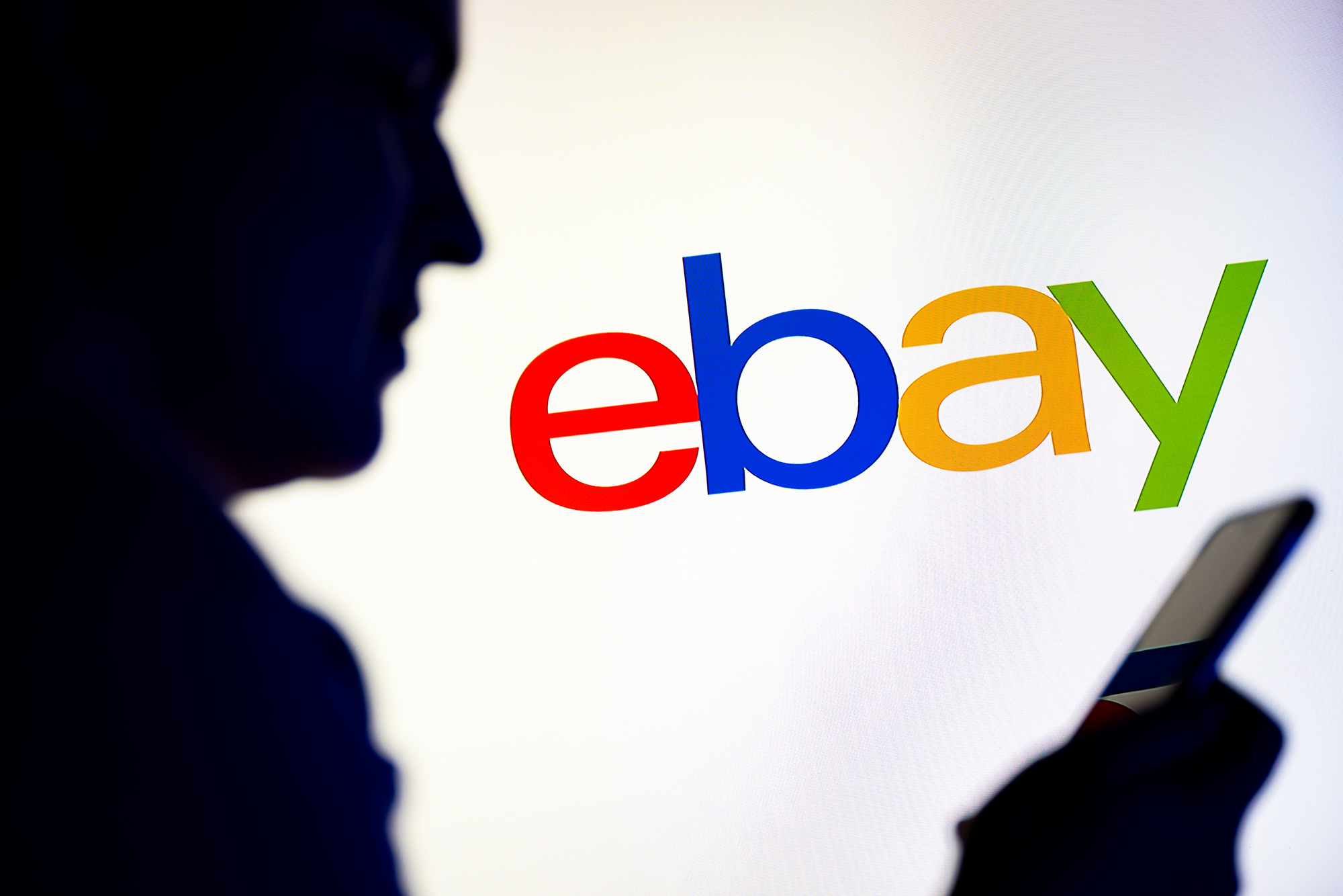 In this photo illustration, a silhouetted individual is seen holding a mobile phone with the eBay logo displayed in the background.
